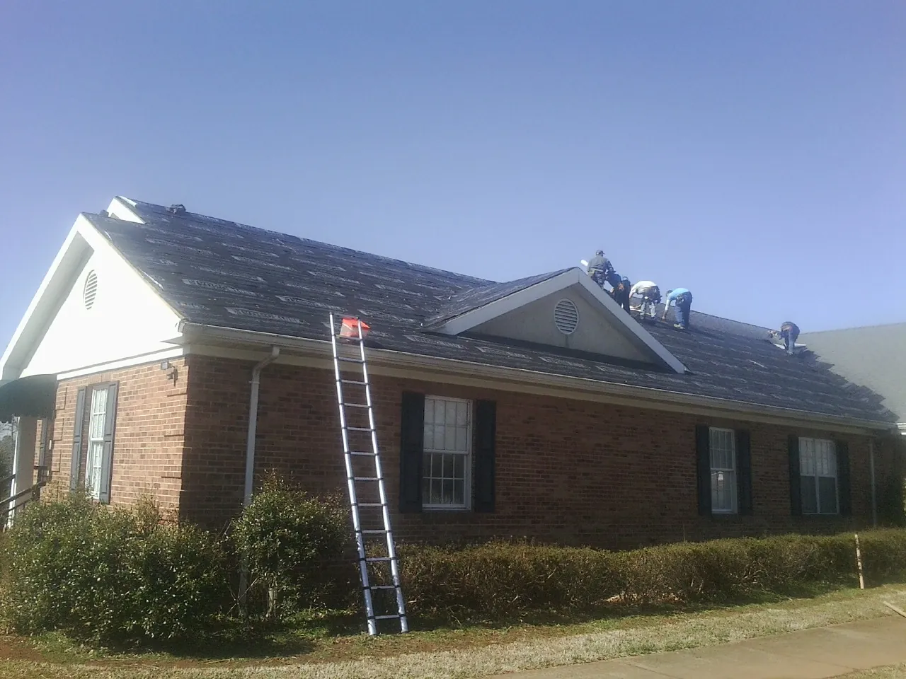 Crew tearing shingles off a brick ranch roof with a ladder set — Metal Roof Repair work by our crew in Holbrook
