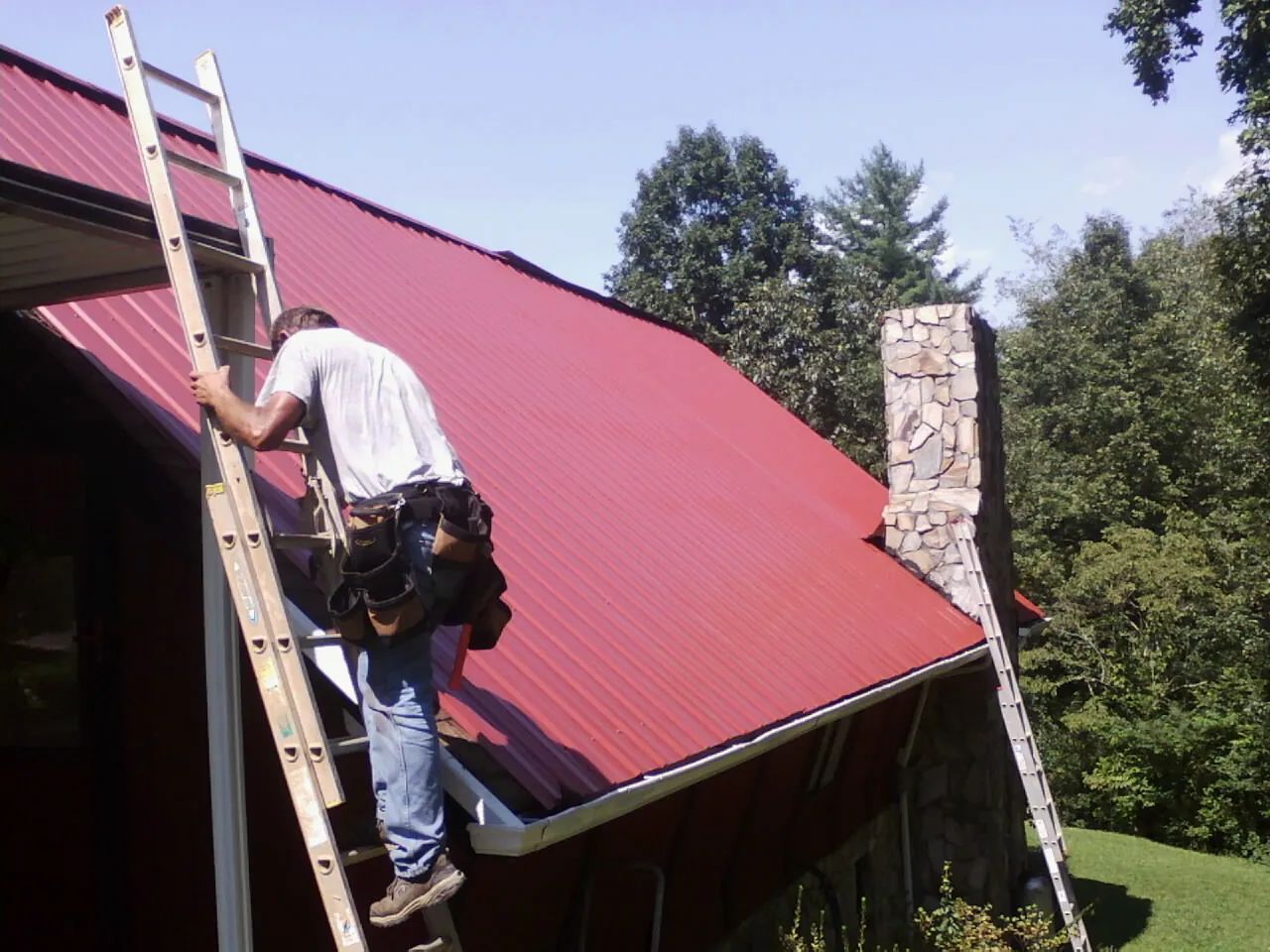 Roofer on a ladder beside a red metal roof and stone chimney — Soffit & Fascia Repair by Roofing Brothers in Holbrook